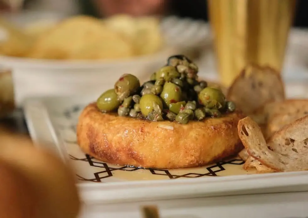 Queso dorado con aceitunas verdes y alcaparras, acompañado de rebanadas de pan en Lilou.